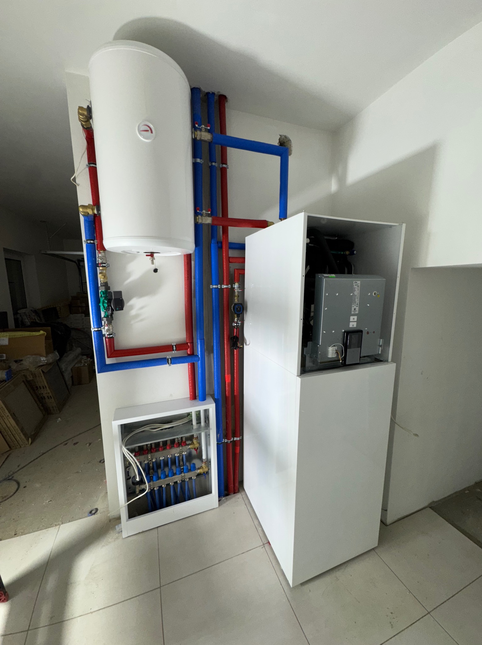Biały zasobnik ciepłej wody, piec w obudowie i rozdzielacz ogrzewania podłogowego z rurami w kolorach niebieskim i czerwonym, zamontowane w pomieszczeniu.