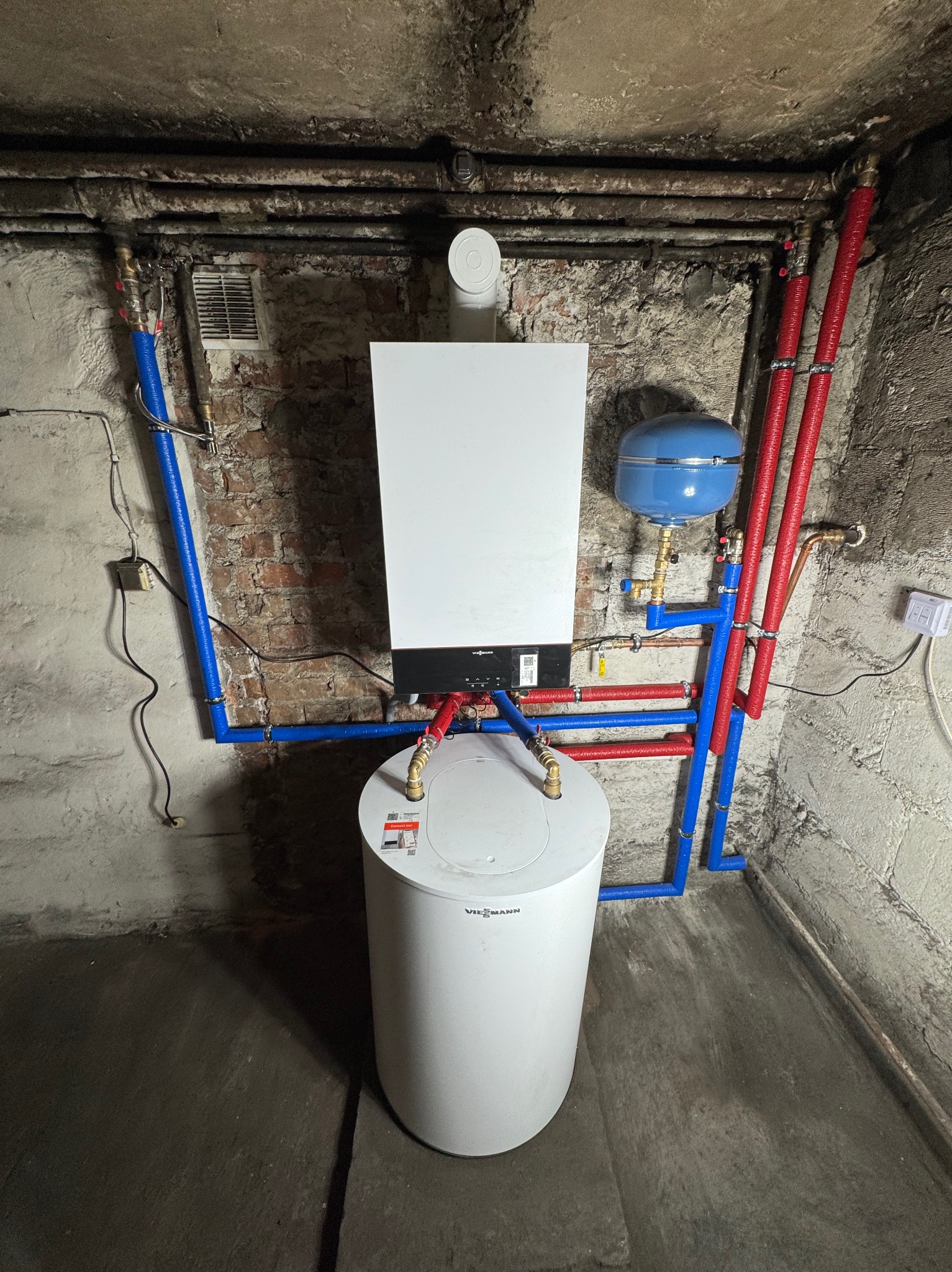 Nowoczesny piec gazowy Viessmann z zasobnikiem i instalacją w piwnicy o surowych ścianach. Rury w kolorach niebieskim i czerwonym. Widoczne elementy instalacji.