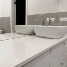 Biały blat łazienkowy z dwiema owalnymi umywalkami nablatowymi i chromowanymi bateriami, odbicie w lustrze ukazuje wannę i wzorzystą tapetę w tle.