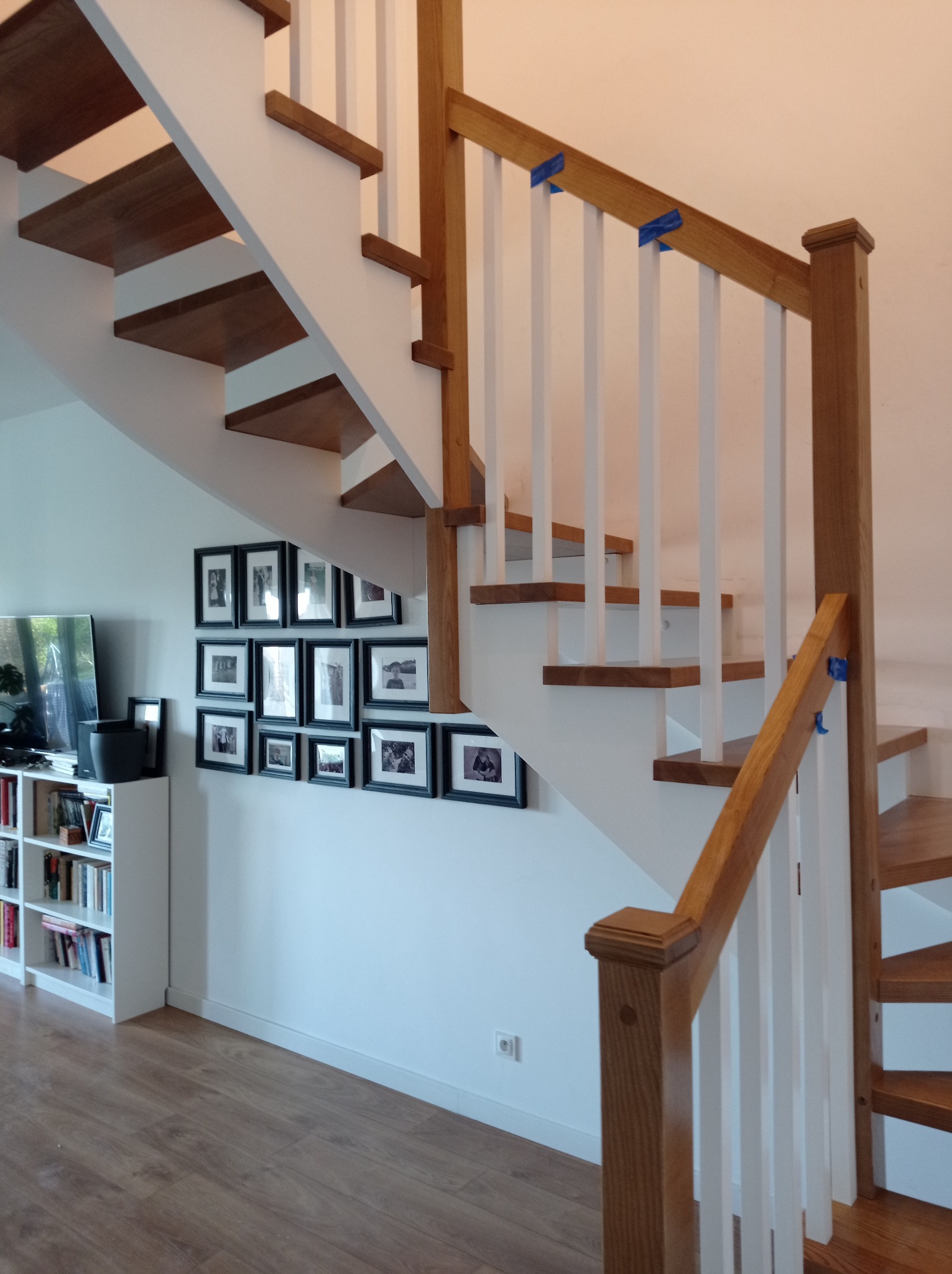 Nowoczesne schody z drewnianymi stopniami i białą konstrukcją, zabezpieczone niebieską taśmą malarską na balustradzie, w tle ściana z galerii czarno-białych zdjęć.