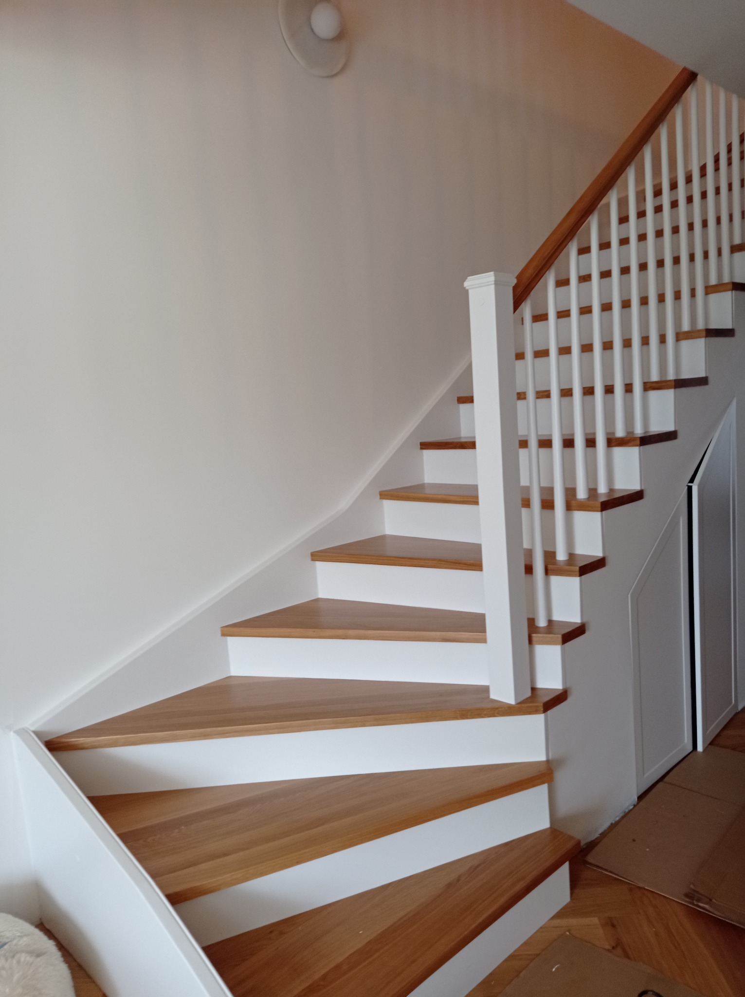 Drewniane schody z białymi podstopnicami i balustradą, widoczne z dołu, z fragmentem parkietu i kartonów na podłodze.