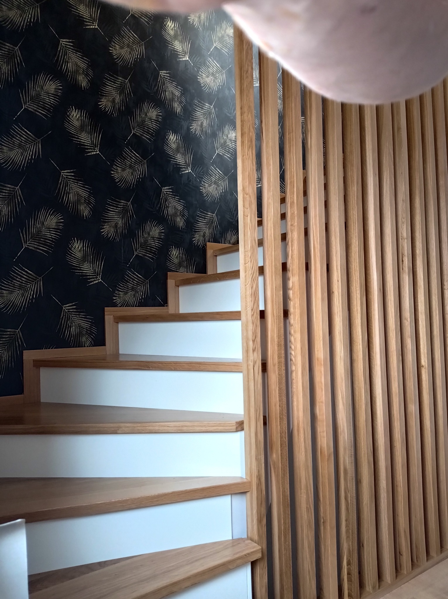 Nowoczesne schody z drewnianymi stopniami i białymi podstopnicami, oddzielone od ściany dekoracyjną, ażurową ścianką z pionowych, drewnianych listew na tle ciemnej ściany z motywem złotych liści.