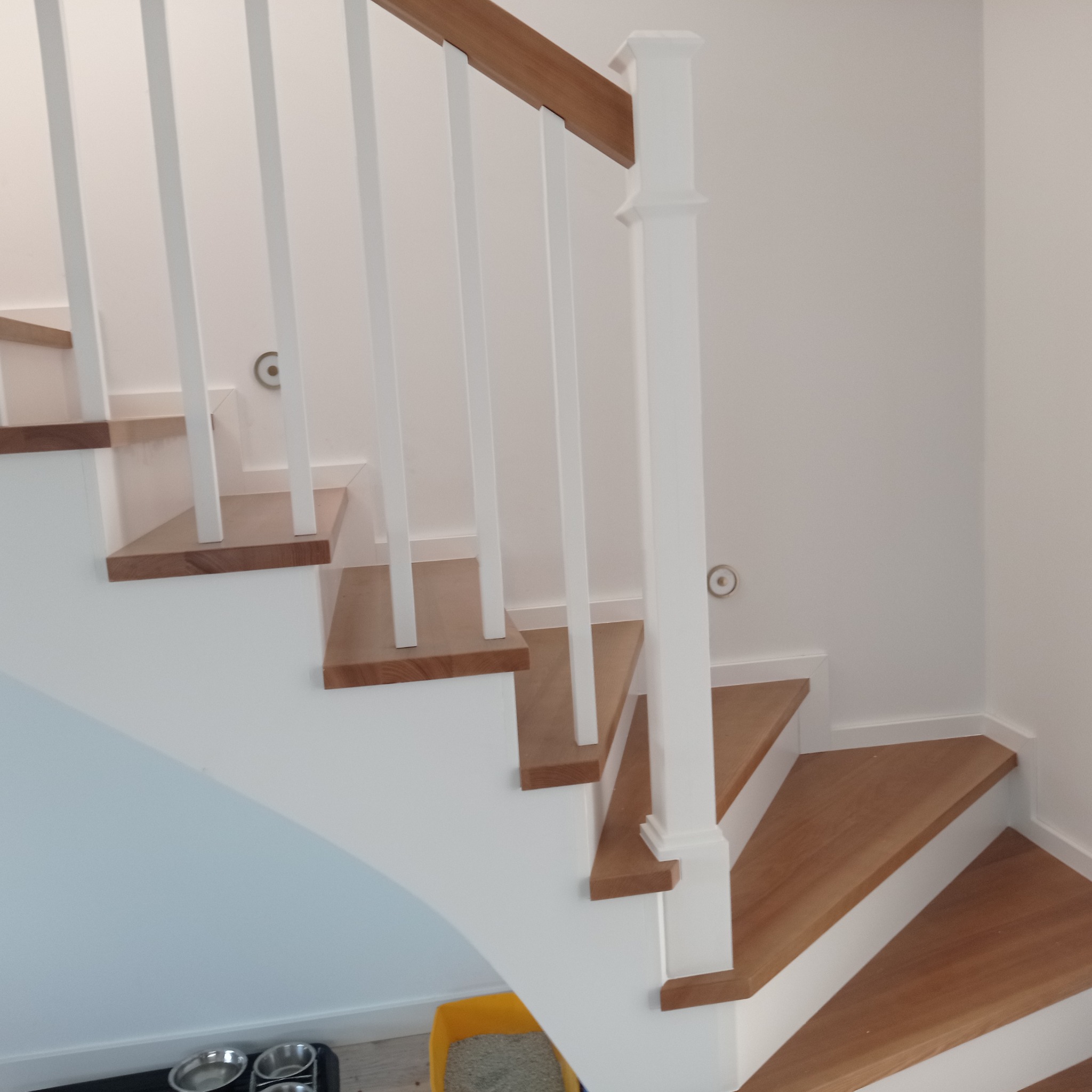 Wnętrze domu z drewnianymi schodami o jasnych stopniach i białych balustradach, widoczne dwa metalowe psie miski i żółty pojemnik na podłodze.