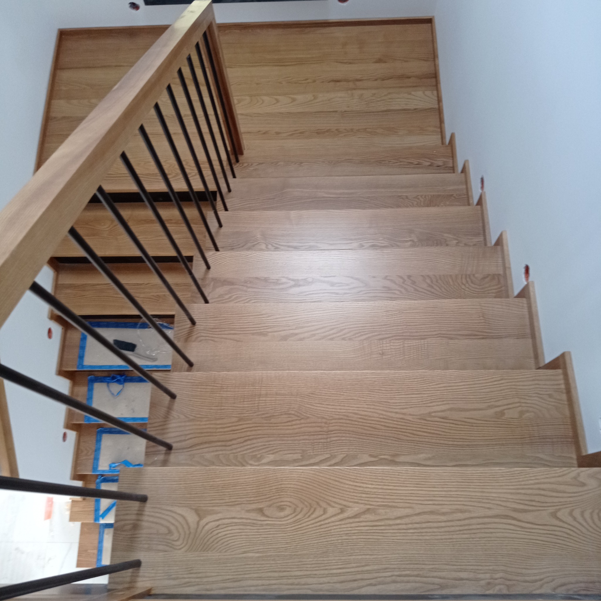 Widok z góry na jasne, drewniane schody z czarnymi, metalowymi prętami balustrady, częściowo zabezpieczone folią malarską.