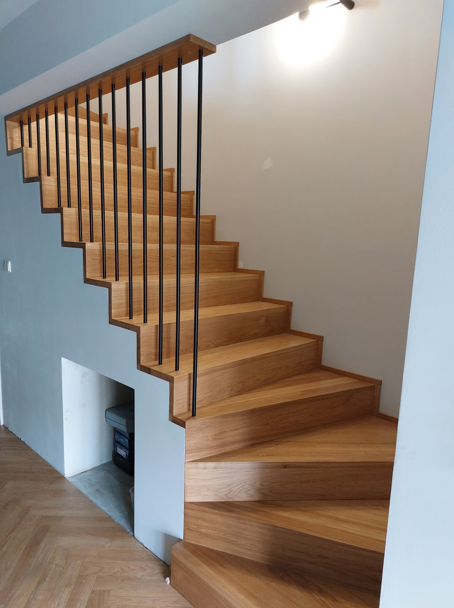 Nowoczesne schody z jasnego drewna z czarnymi, pionowymi balustradami, widoczne z dołu, z zagospodarowaną przestrzenią pod schodami.