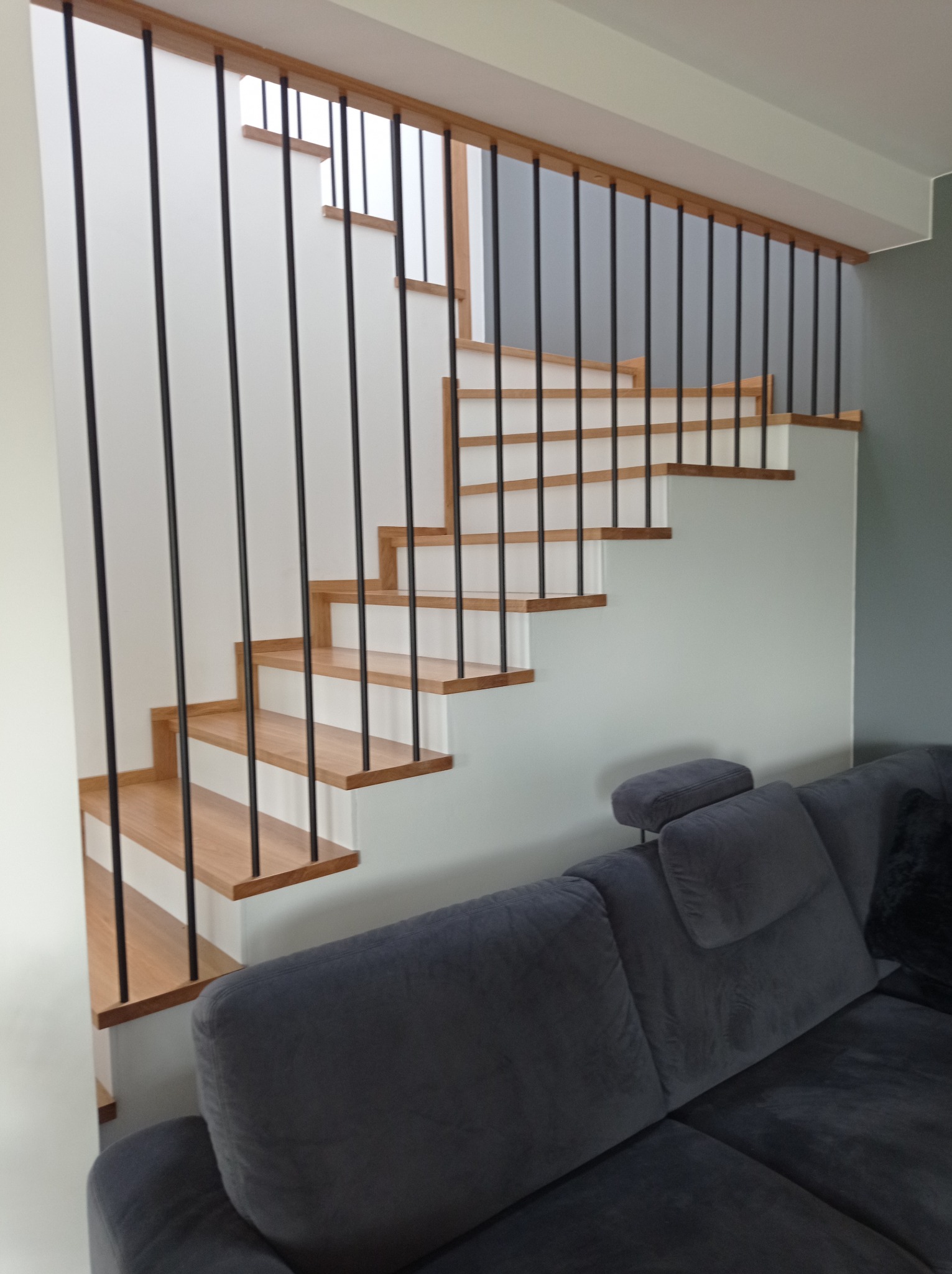Drewniane schody z czarnymi, metalowymi balustradami w nowoczesnym wnętrzu, częściowo zasłonięte przez szarą sofę.