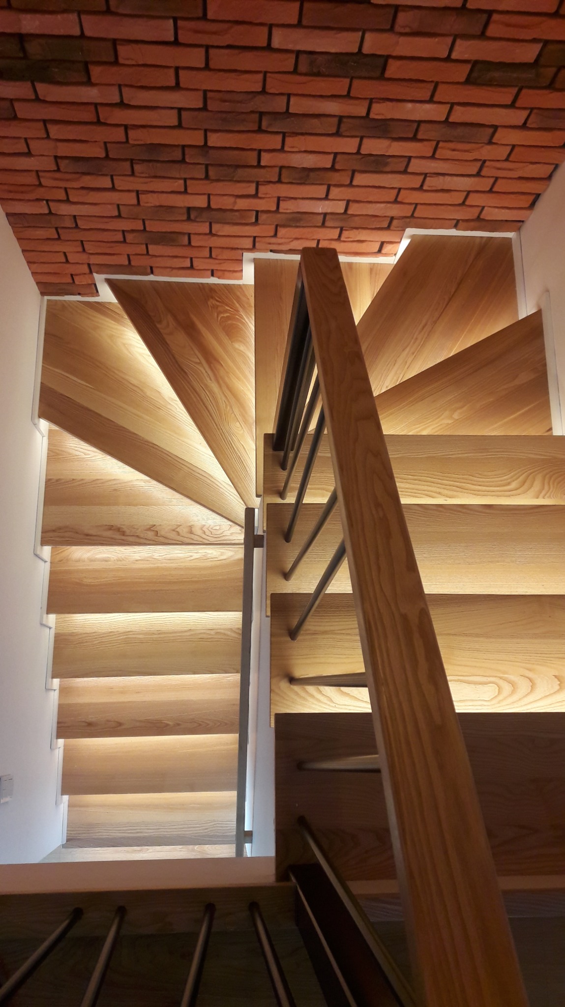 Kaskadowe stopnie z jasnego drewna z podświetleniem LED, widok z góry, z balustradą z ciemnego metalu i drewnianą poręczą, ściana z cegły na górze.