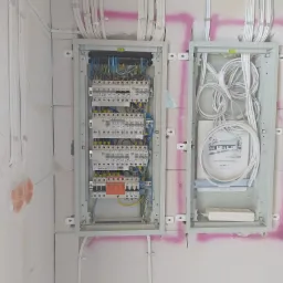 Dwa panele elektryczne zamontowane na ścianie z betonu kom&oacute;rkowego. W lewym panelu widoczne bezpieczniki i okablowanie. W prawym panelu zwinięte kable i dokumentacja techniczna projektu domu 'Mokka...