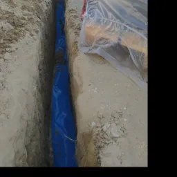 Wykop w ziemi wyłożony niebieską folią, prawdopodobnie przygotowany pod przyłącze elektryczne, częściowo zakryty przez przezroczystą folię ochronną.