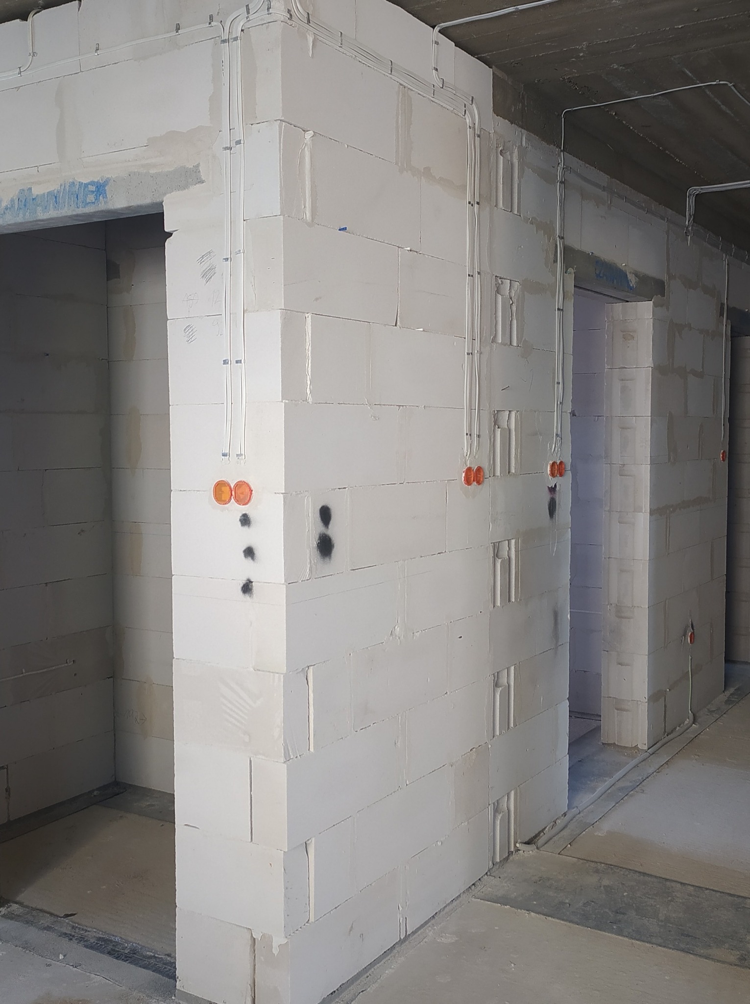 Surowy stan: białe ściany z betonu komórkowego z widocznymi przewodami elektrycznymi w peszlach, pomarańczowe puszki instalacyjne podtynkowy zamontowane, widoczne otwory drzwiowe, szara betonowa...