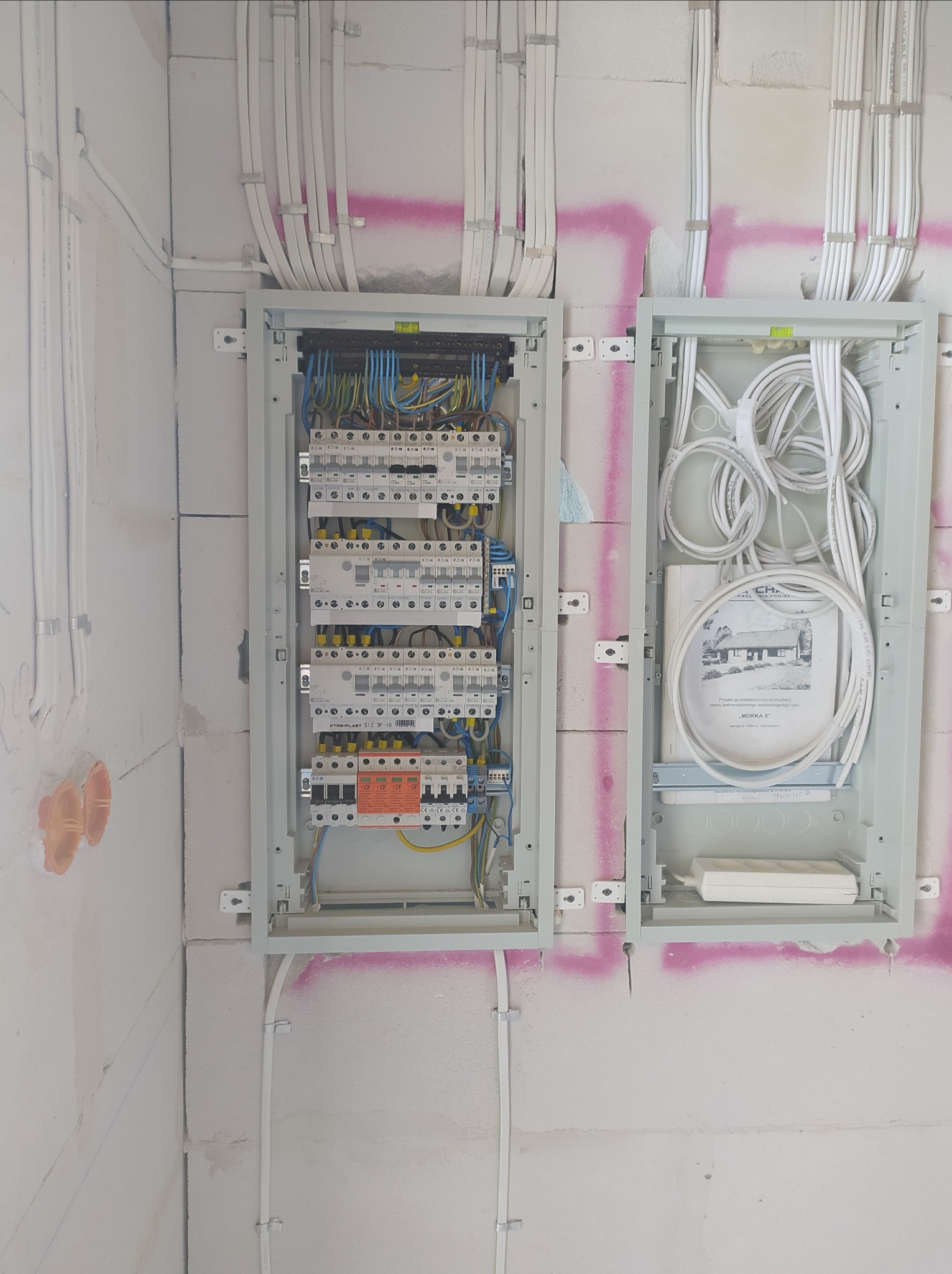 Dwa panele elektryczne zamontowane na ścianie z betonu komórkowego. W lewym panelu widoczne bezpieczniki i okablowanie. W prawym panelu zwinięte kable i dokumentacja techniczna projektu domu 'Mokka...