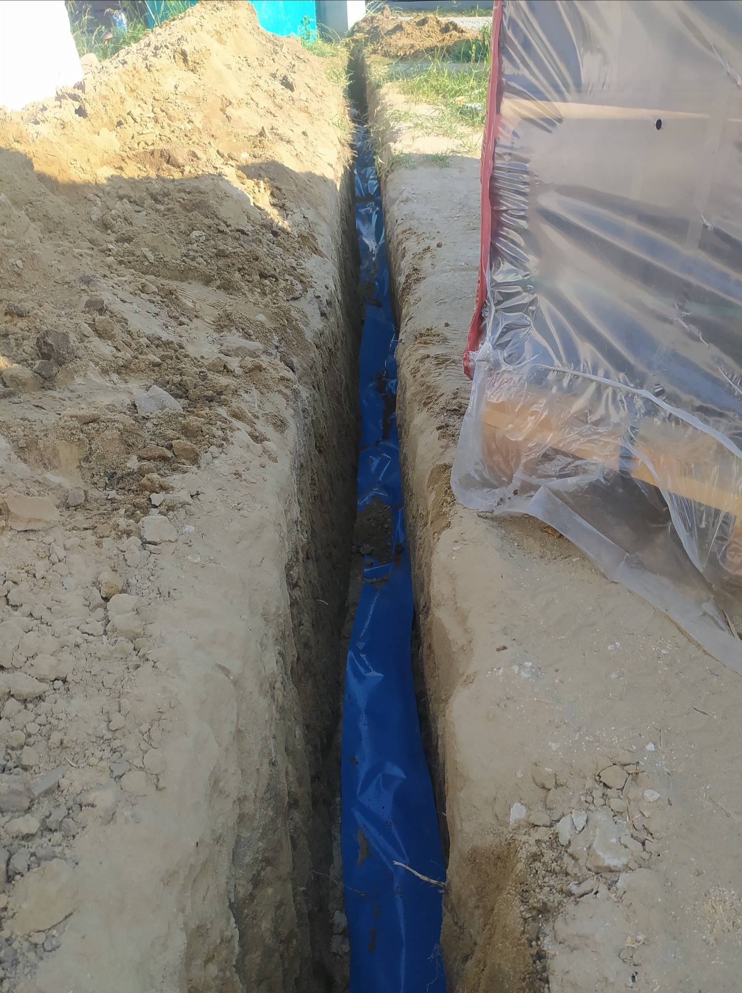 Wykopany wąski rów w ziemi, wyłożony niebieską folią, prawdopodobnie pod kable elektryczne, z widocznym fragmentem maszyny w tle, przykrytej folią ochronną.