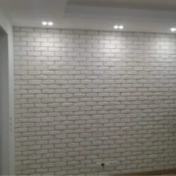 Ściana wewnętrzna wykończona dekoracyjną cegłą pomalowaną na biało, z widocznymi spoinami, oświetlona punktowymi lampami sufitowymi.