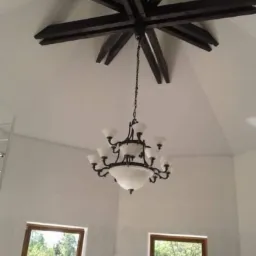 Wysokie pomieszczenie z gładkimi, białymi ścianami, drewnianą dekoracją sufitową w kształcie gwiazdy i żyrandolem. Widoczne okna z widokiem na zieleń oraz drabina w rogu.