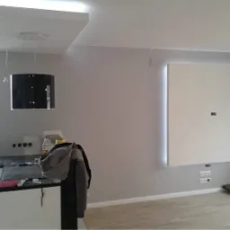 Wnętrze w trakcie wykańczania: szara ściana, zabudowa telewizyjna z podświetleniem LED, fragment kuchni z czarnym okapem i niedokończone gniazdka elektryczne.