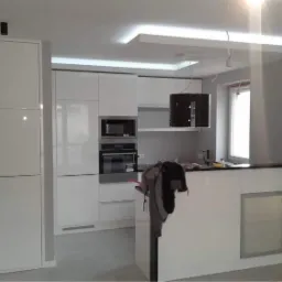 Nowoczesna kuchnia z białymi, lakierowanymi frontami szafek, wyspą z czarnym blatem i oświetleniem LED pod sufitem, ściany pomalowane na szaro.