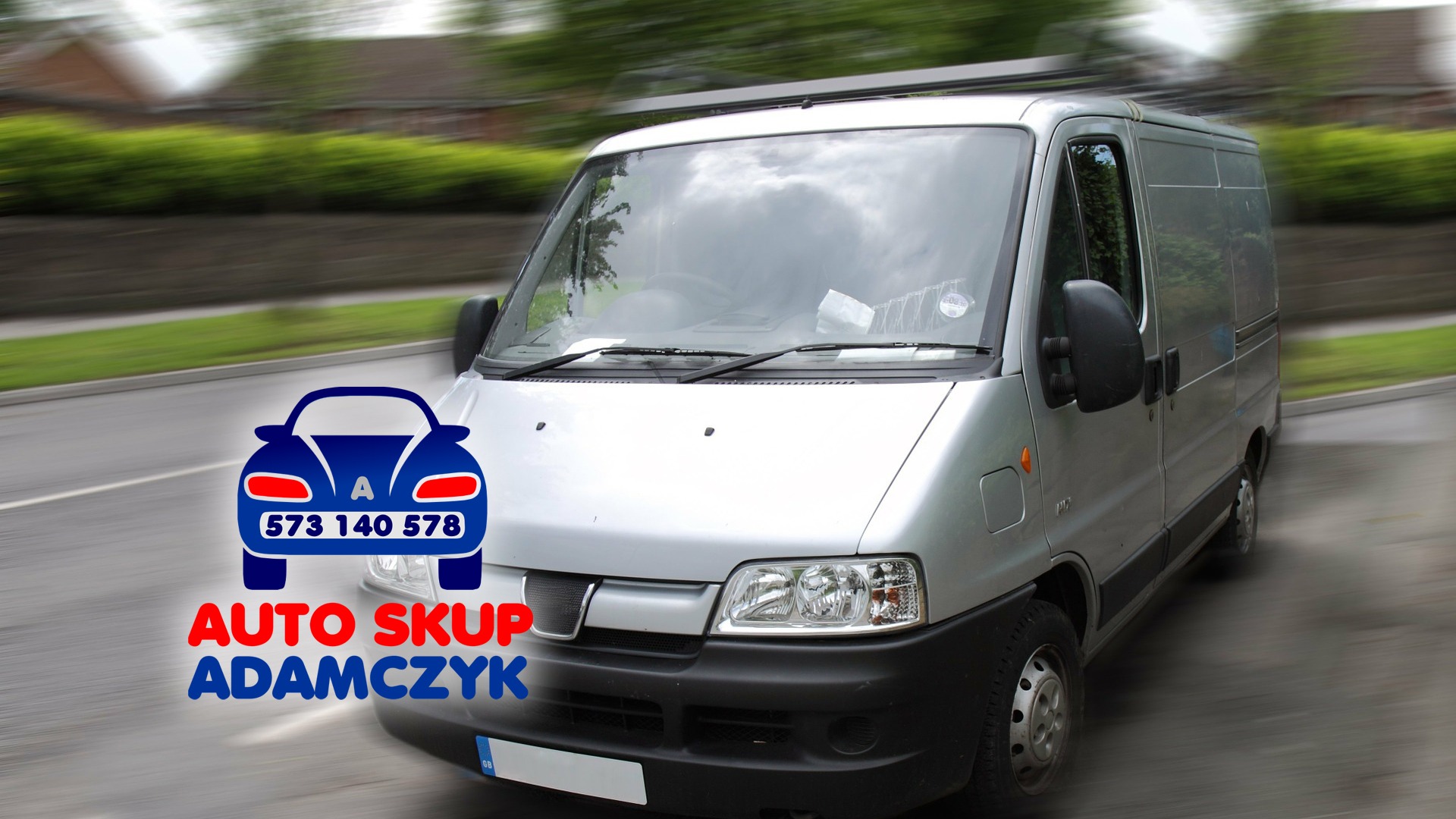 Srebrny bus dostawczy z grafiką 'Auto Skup Adamczyk' i numerem telefonu, sfotografowany w ruchu na tle rozmytej zieleni.