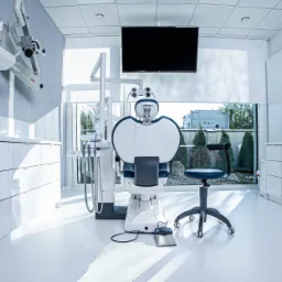 Jasne, nowoczesne wnętrze gabinetu lekarskiego z białym fotelem dentystycznym, narzędziami i krzesłem na kółkach, widok na zieleń za oknem.