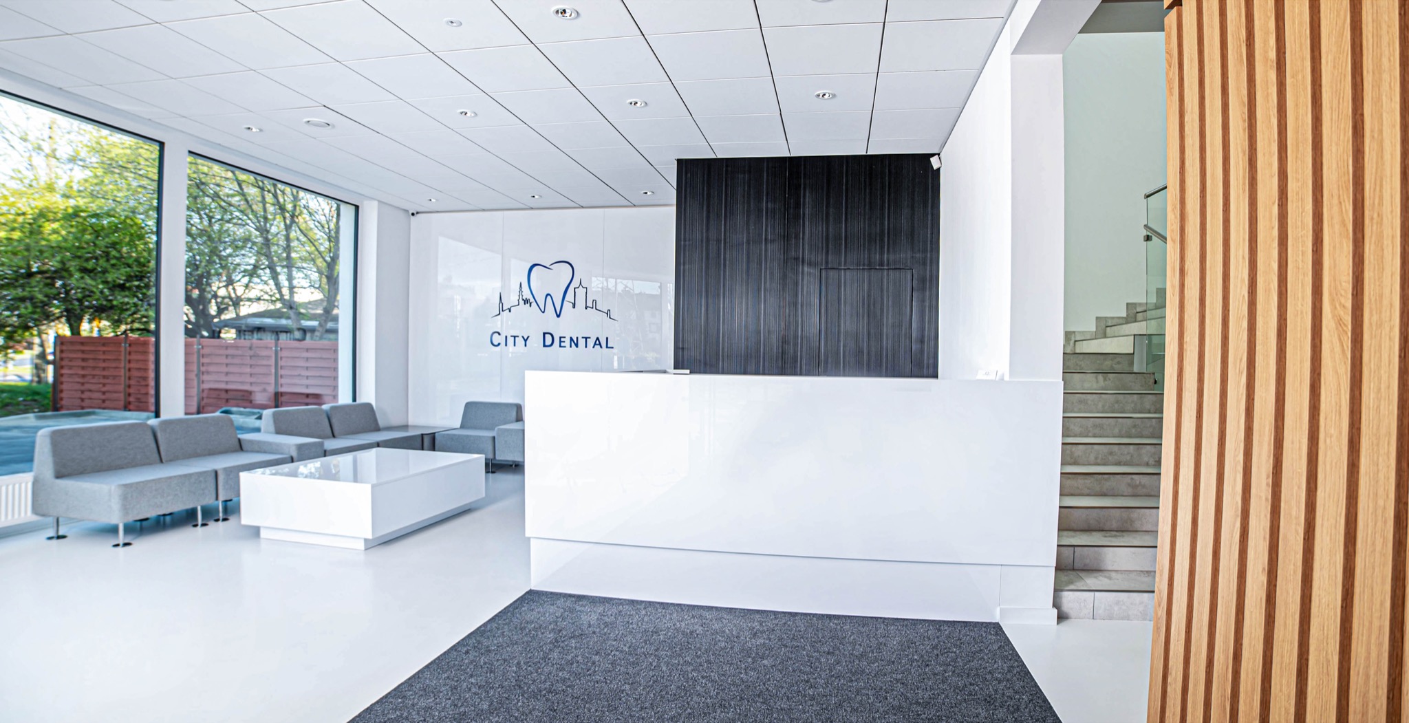 Jasne, nowoczesne wnętrze kliniki dentystycznej z recepcją, szarą poczekalnią i logo City Dental na szklanej ściance.