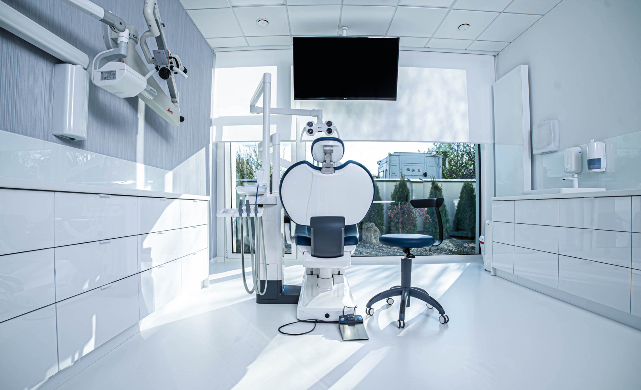 Jasne, nowoczesne wnętrze gabinetu lekarskiego z białym fotelem dentystycznym, narzędziami i krzesłem na kółkach, widok na zieleń za oknem.