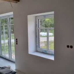 Wnętrze w trakcie prac wykończeniowych: ściany po nałożeniu gładzi, z widocznymi oknami PCV, drewnianym stropem i puszkami elektrycznymi bez osprzętu.