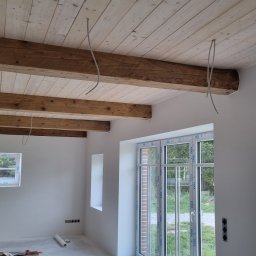 MAXDOM Roman KOEPPEN - Wnętrze pokoju z drewnianym stropem belkowym, świeżo pomalowanymi ścianami i dużymi oknami, widoczne narzędzia malarskie na podłodze.