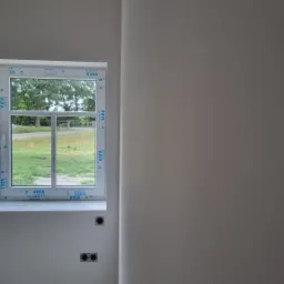 Ściana po szpachlowaniu i malowaniu na szaro, widoczne gniazdka elektryczne i okno z widokiem na zieleń.