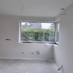 Pokój w trakcie wykańczania: jedna ściana z surową fakturą, pozostałe białe, okno z widokiem na zieleń, widoczne instalacje elektryczne i hydrauliczne.