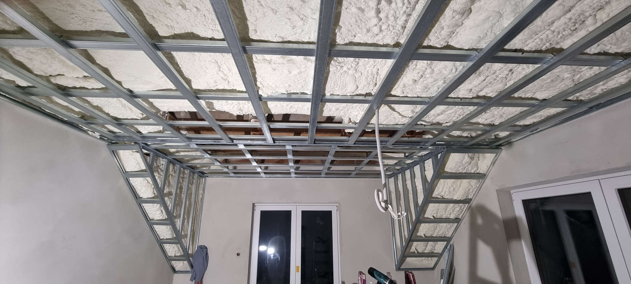 Sufit podwieszany w trakcie montażu: metalowa konstrukcja wypełniona pianką izolacyjną, widoczne kable i fragment okna.