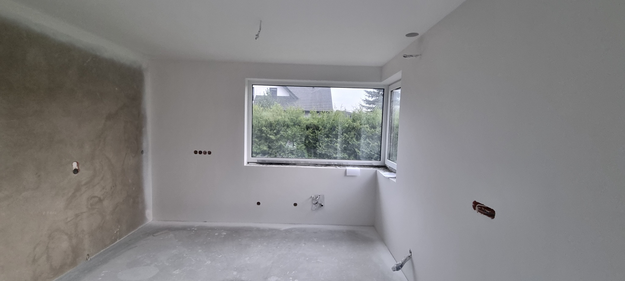 Pokój w trakcie wykańczania: jedna ściana z surową fakturą, pozostałe białe, okno z widokiem na zieleń, widoczne instalacje elektryczne i hydrauliczne.