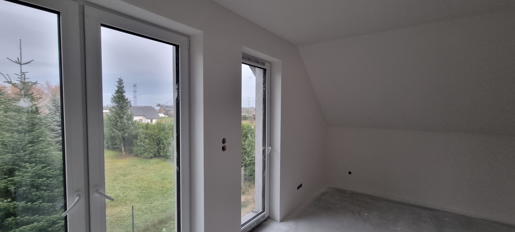 Wnętrze poddasza z dwoma oknami PCV, ściany i sufit świeżo wykończone na biało, widoczne ślady po pracach wykończeniowych na podłodze.