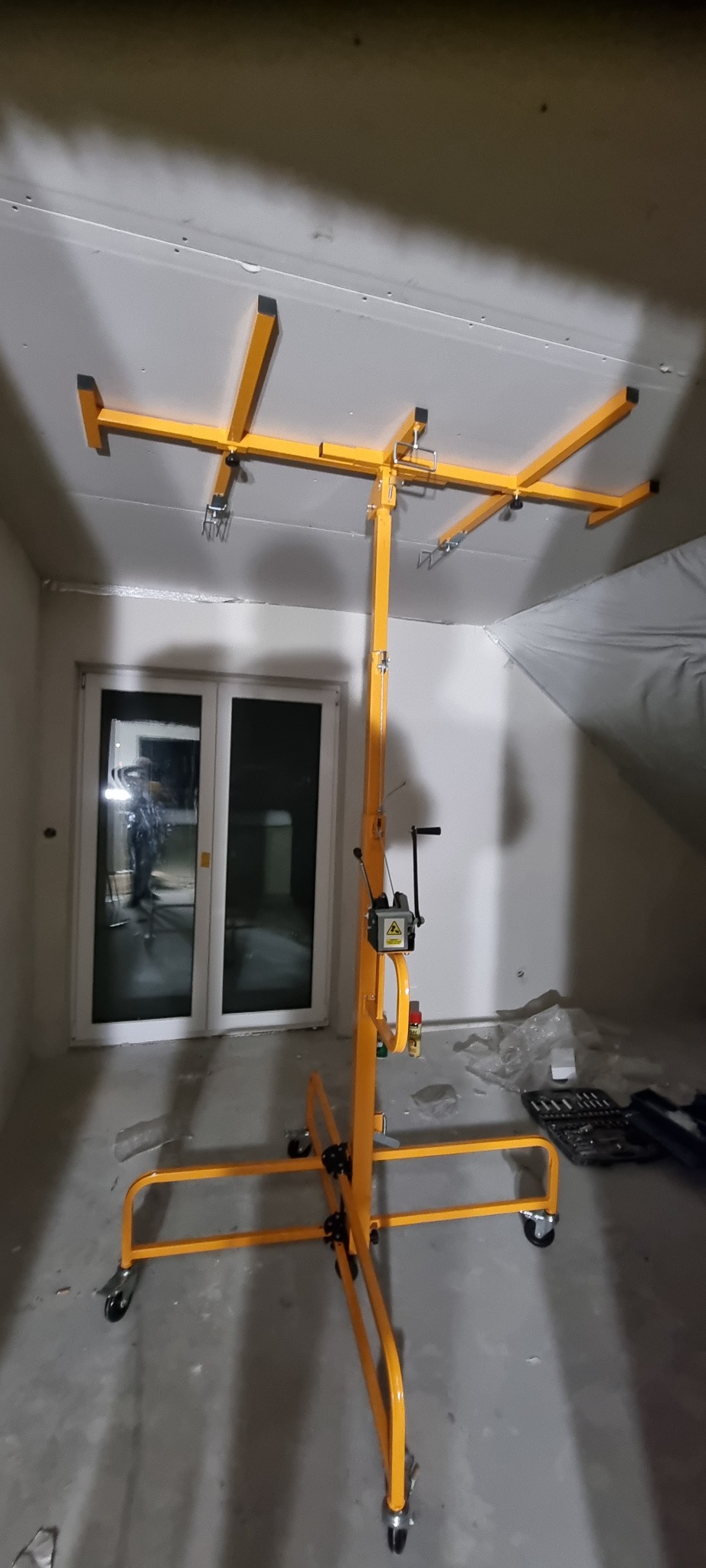 Żółty podnośnik do płyt gipsowo-kartonowych ustawiony w pomieszczeniu z częściowo zamontowanymi płytami na suficie, widoczne narzędzia w tle.