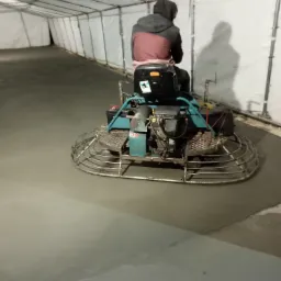 Pracownik w ciemnej bluzie siedzi na maszynie do zacierania betonu wewnątrz namiotu, wyrównując świeżo wylaną posadzkę.