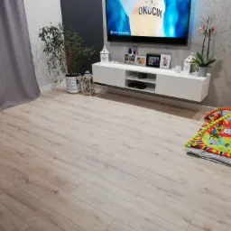 Jasne panele podłogowe w salonie z białym, wiszącym stolikiem RTV, telewizorem wyświetlającym reklamę piwa, roślinami doniczkowymi i kolorową matą edukacyjną dla dziecka.