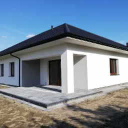 Nowoczesny, parterowy dom z białymi ścianami, czarnym dachem i szarą kostką brukową na tarasie, sfotografowany w słoneczny dzień.