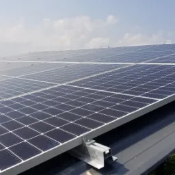 Panele fotowoltaiczne zamontowane na dachu z widocznymi elementami mocującymi i fragmentem sylwetki instalatora w prawym dolnym rogu.