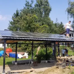 Montaż paneli fotowoltaicznych na metalowej konstrukcji w ogrodzie, dwóch mężczyzn pracuje na dachu, widoczne drzewa i zabudowania w tle.