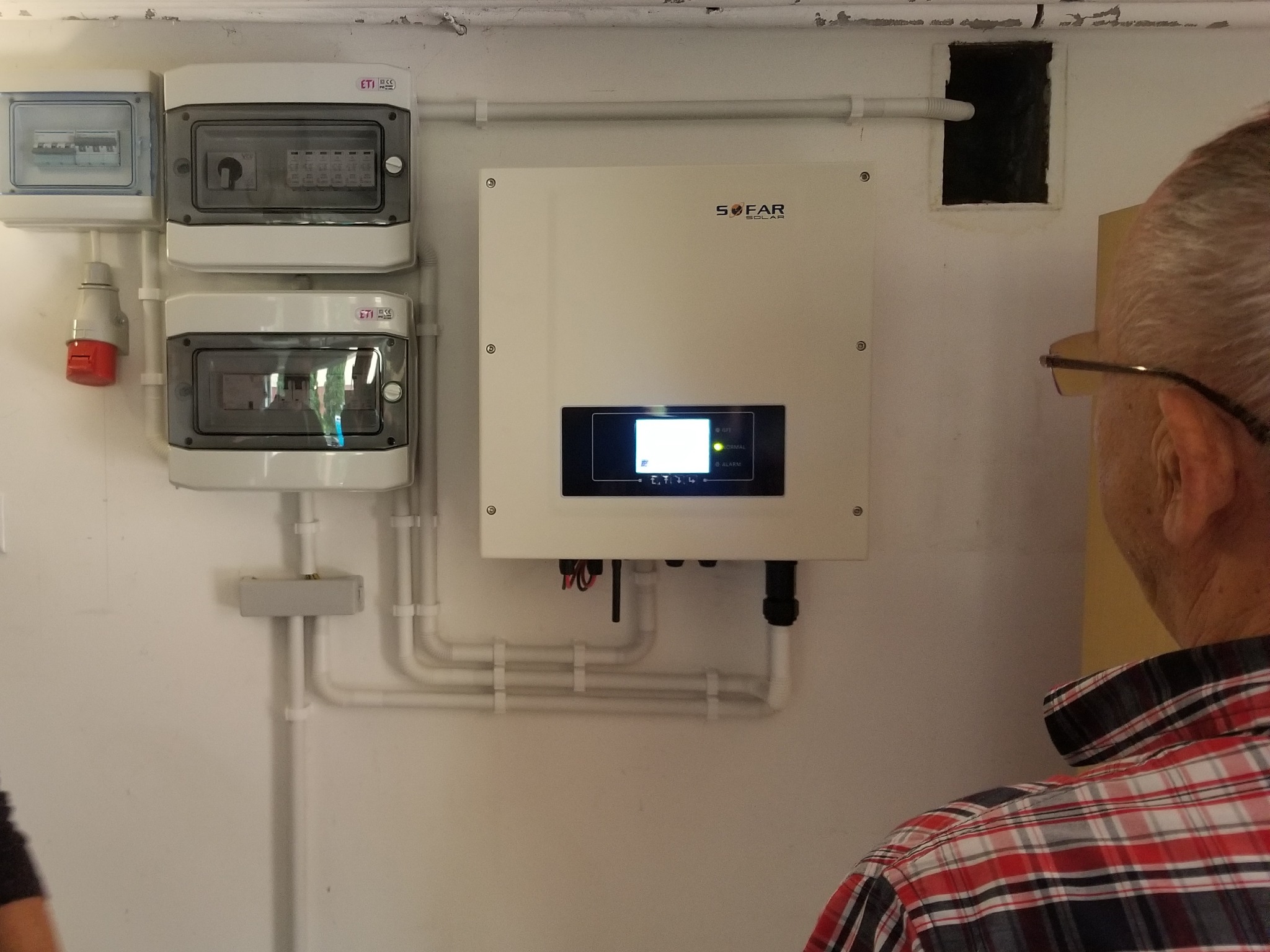 Instalacja paneli fotowoltaicznych: biała skrzynka inwertera Sofar Solar zamontowana na ścianie obok skrzynek rozdzielczych, widoczny fragment osoby w okularach i koszuli w kratę.