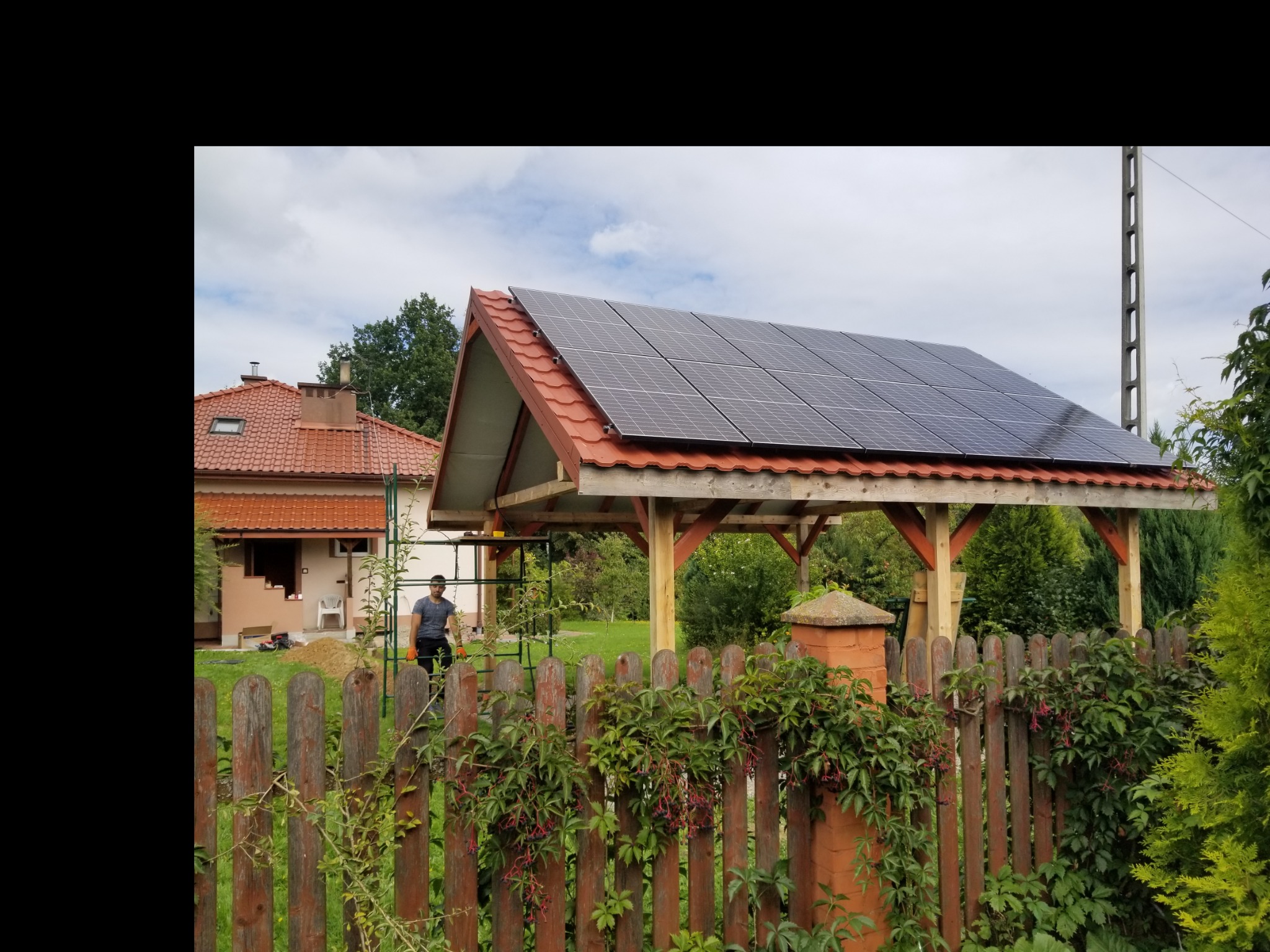 Montaż paneli słonecznych na drewnianej konstrukcji w Gorlicach, widoczne elementy instalacji i pracownik w tle, ogrodzenie z roślinnością na pierwszym planie.