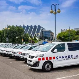 Szereg białych samochodów firmy ochroniarskiej Karabela Ochrona zaparkowanych na parkingu z budynkiem w tle pod błękitnym niebem.