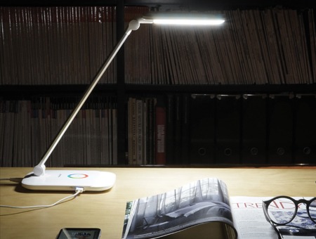 Biała lampka biurkowa LED z panelem dotykowym RGB, ustawiona na biurku z magazynem i okularami na tle regału z książkami.