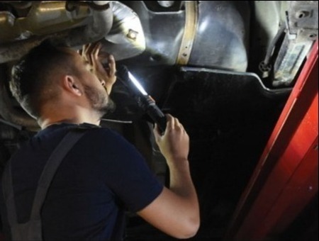 Mechanik w kombinezonie roboczym sprawdza podwozie samochodu za pomocą lampy inspekcyjnej, skupiając się na elementach układu wydechowego i zbiorniku paliwa.