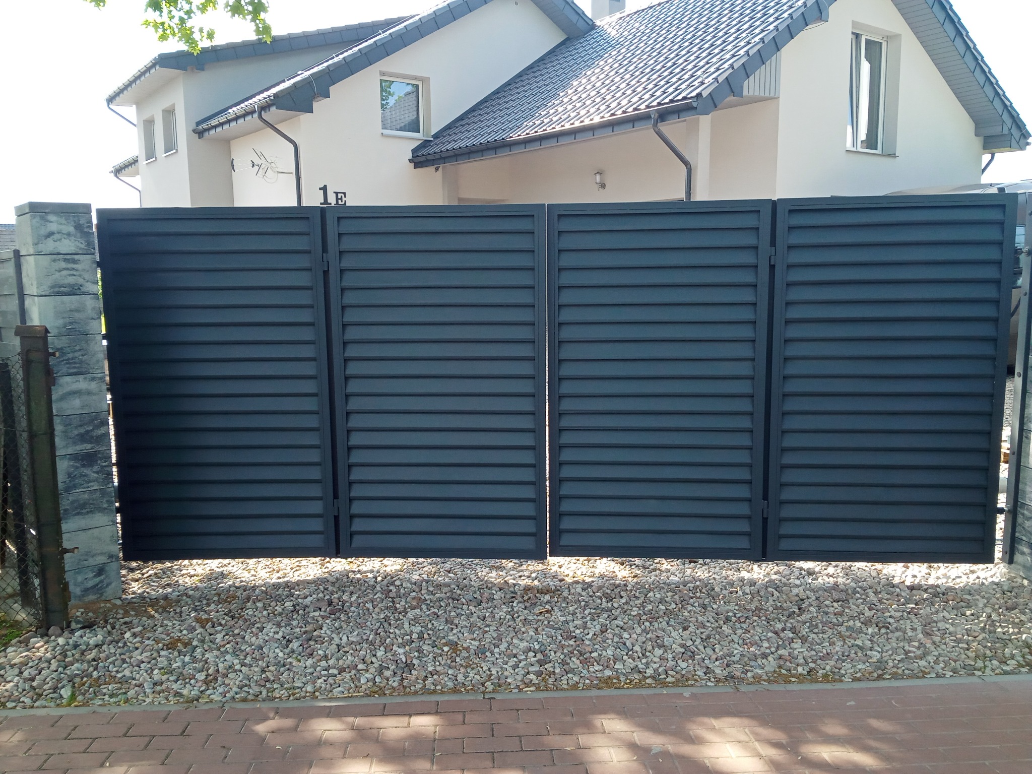 Nowoczesna, pozioma brama wjazdowa w kolorze grafitowym, z widocznym domem w tle i żwirowym podłożem.