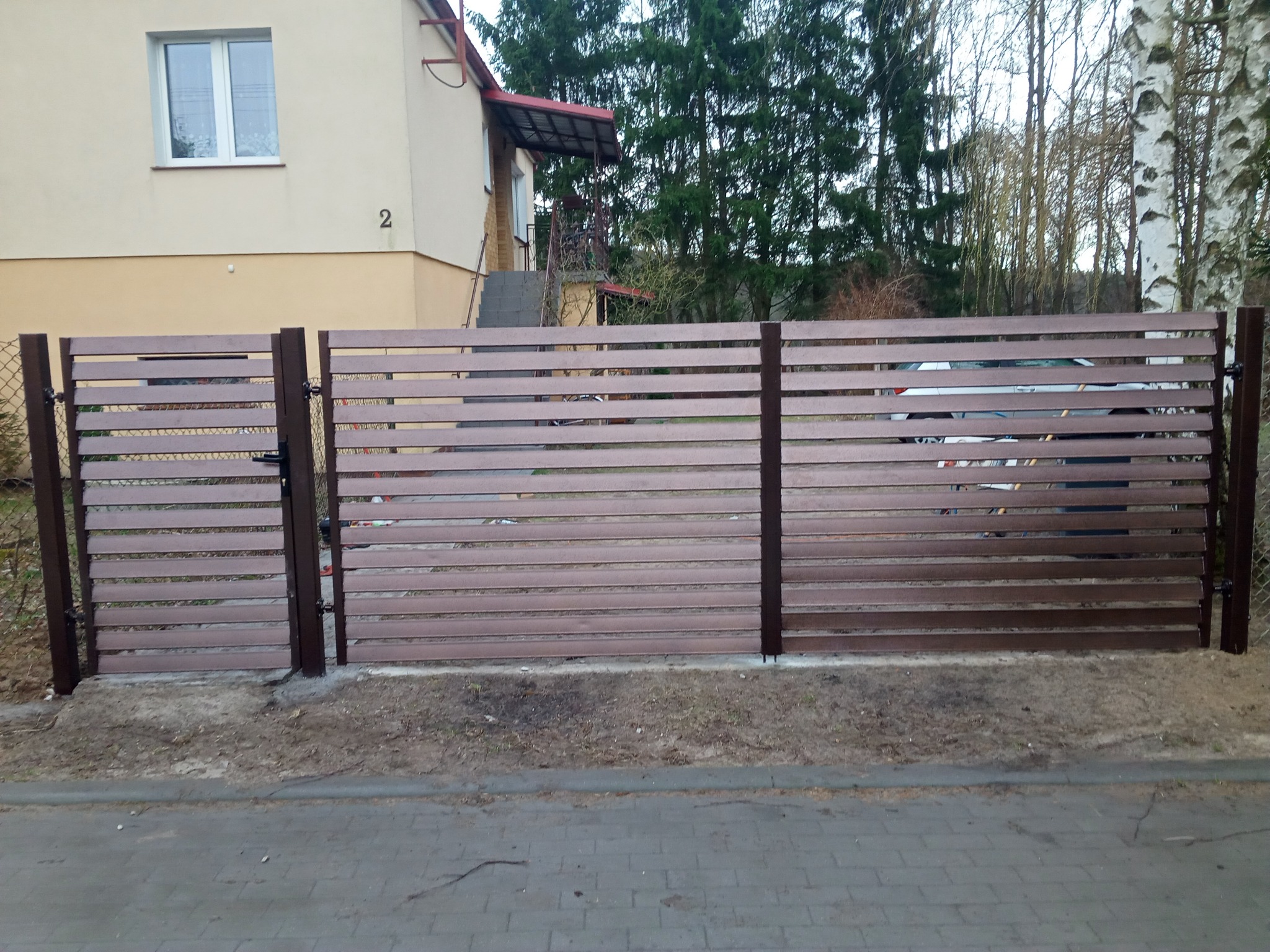 Brązowe ogrodzenie panelowe z poziomych desek, widoczny dom z numerem 2 w tle, brama zamknięta, teren częściowo niezagospodarowany.