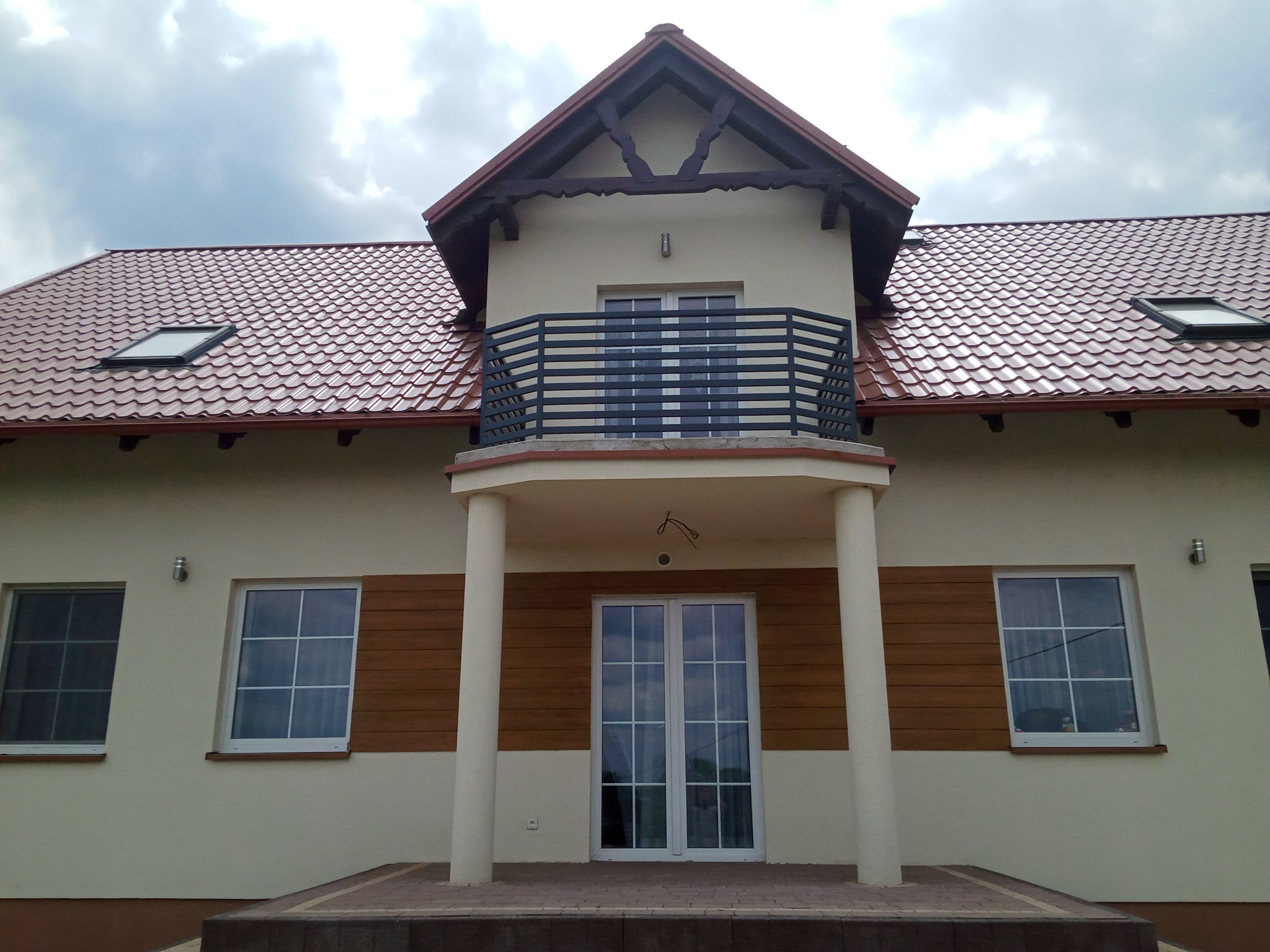 Elewacja domu z balkonem z metalową balustradą, brązową dachówką i drewnianymi panelami dekoracyjnymi.