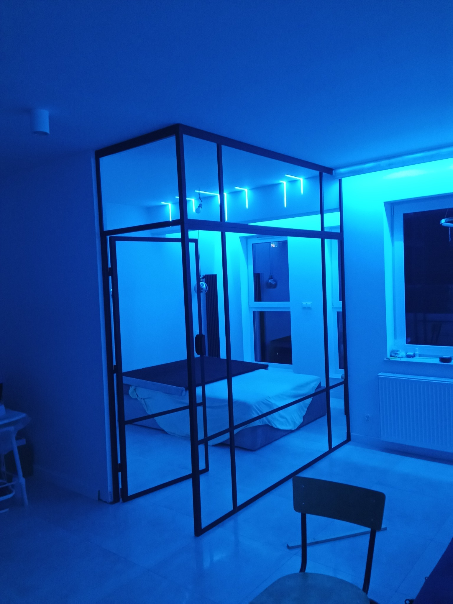 Nowoczesna, czarna konstrukcja stalowa oddzielająca przestrzeń sypialni, oświetlona niebieskim światłem LED, z widocznym łóżkiem i krzesłem w tle.