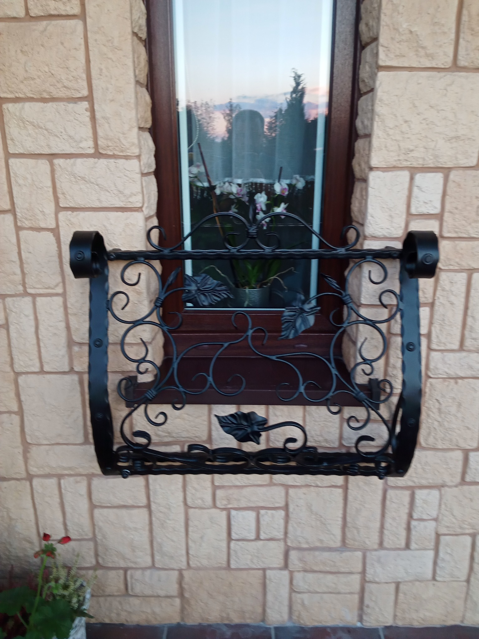 Kuta, czarna balustrada okienna z motywem liści, zamontowana na elewacji z jasnej cegły, z kwiatami widocznymi w oknie.