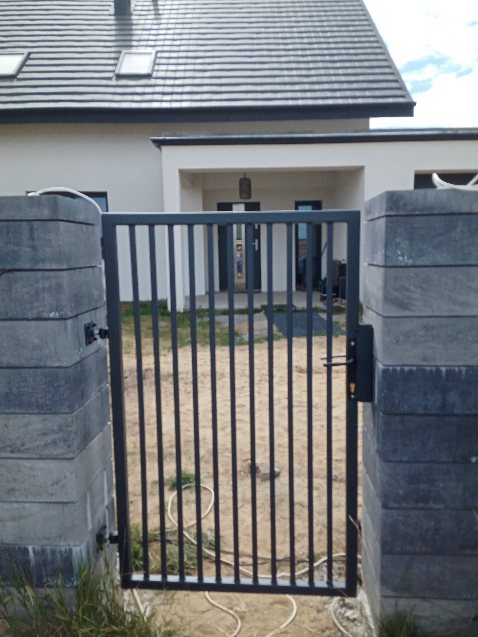 Nowoczesna, grafitowa furtka z pionowych prętów, zamontowana między słupkami z szarej kostki betonowej, na tle domu z ciemną dachówką.