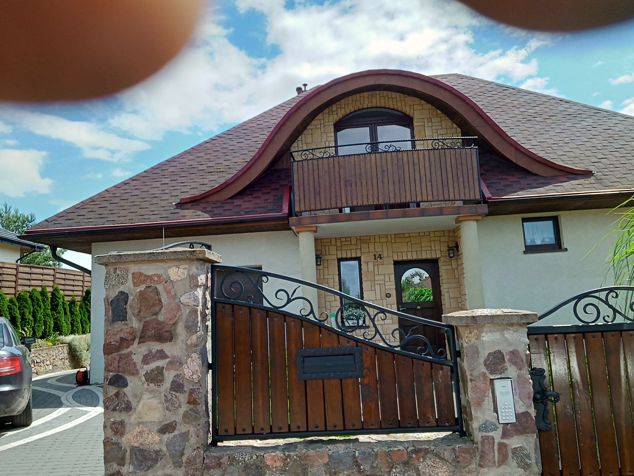 Drewniana brama wjazdowa z metalowymi zdobieniami i kamiennymi słupkami, dopasowana do elewacji domu z balkonem.
