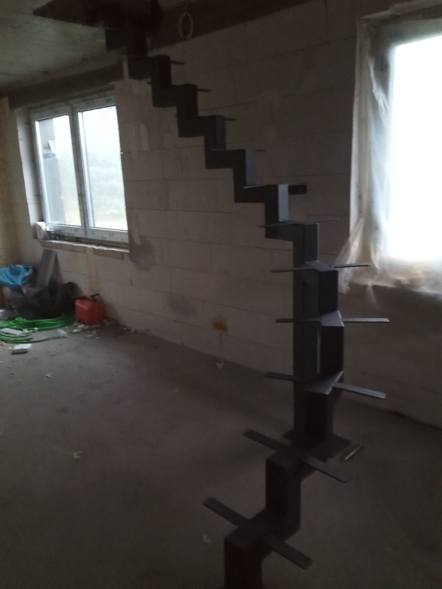 Surowe, metalowe schody bez stopni w stanie surowym, w trakcie montażu wewnątrz budynku, widoczne bloczki betonowe ścian i okno z folią ochronną.